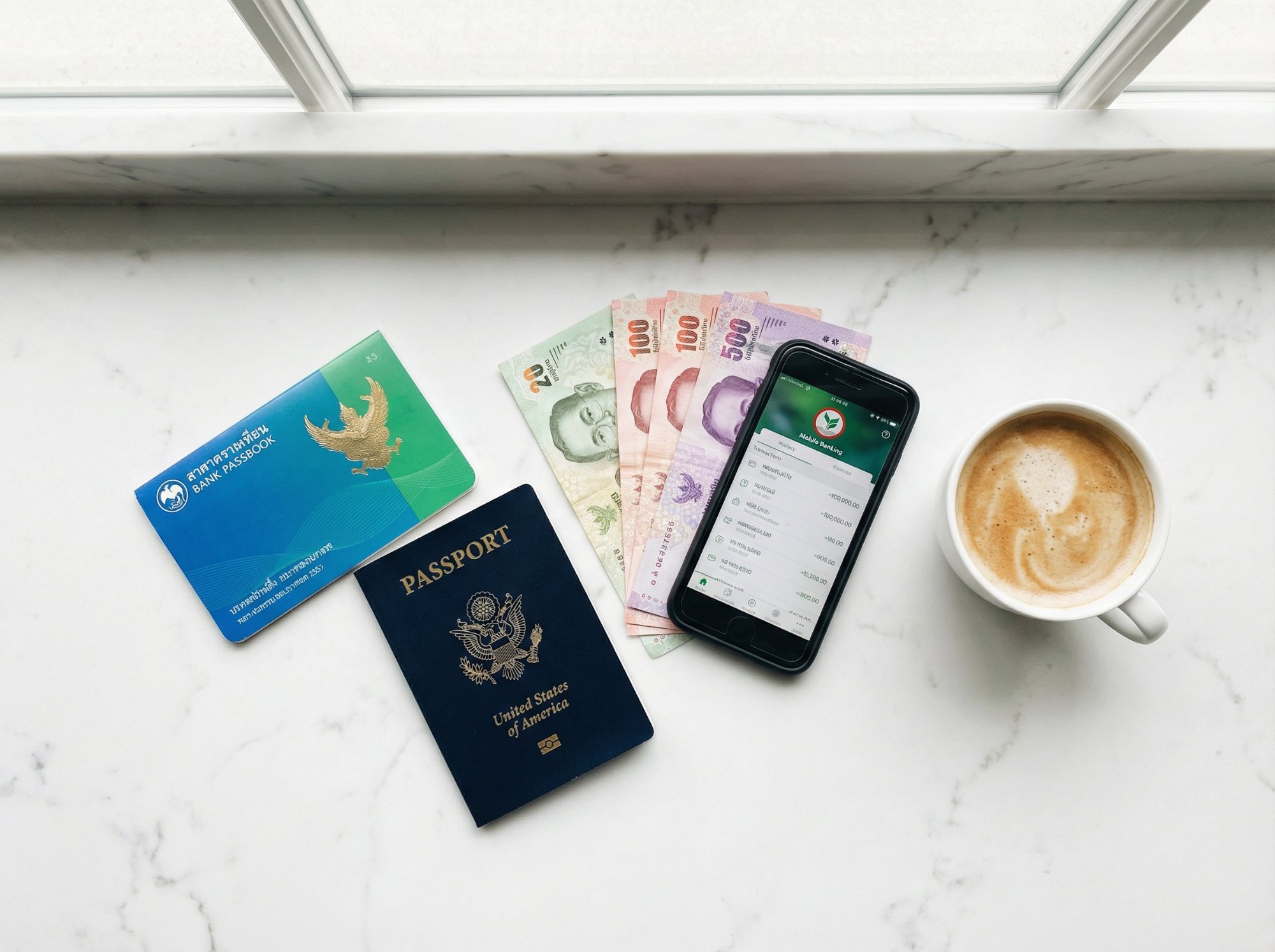 Thai bank passbook, passport, baht bills, mobile banking app and coffee on marble surface